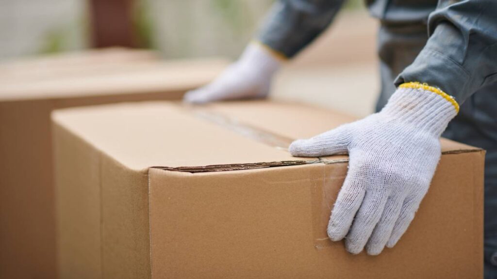 Close-up of a person wearing gloves and carrying a cardboard moving box. - where to get moving boxes