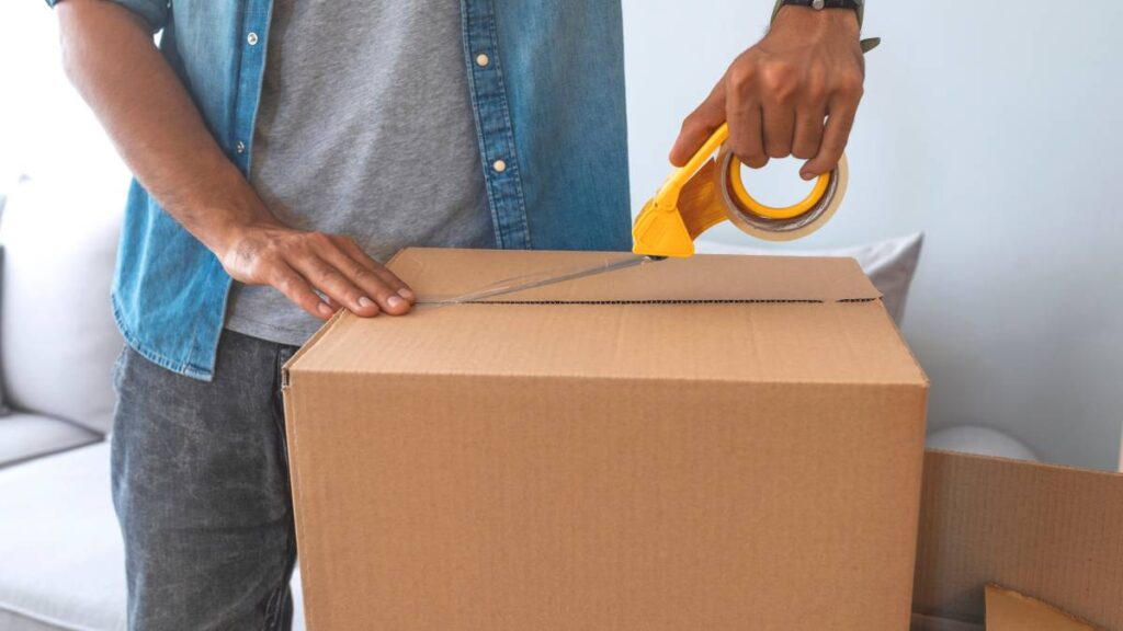Close-up of a person sealing a brown cardboard box with packing tape. - where to get inexpensive moving boxes