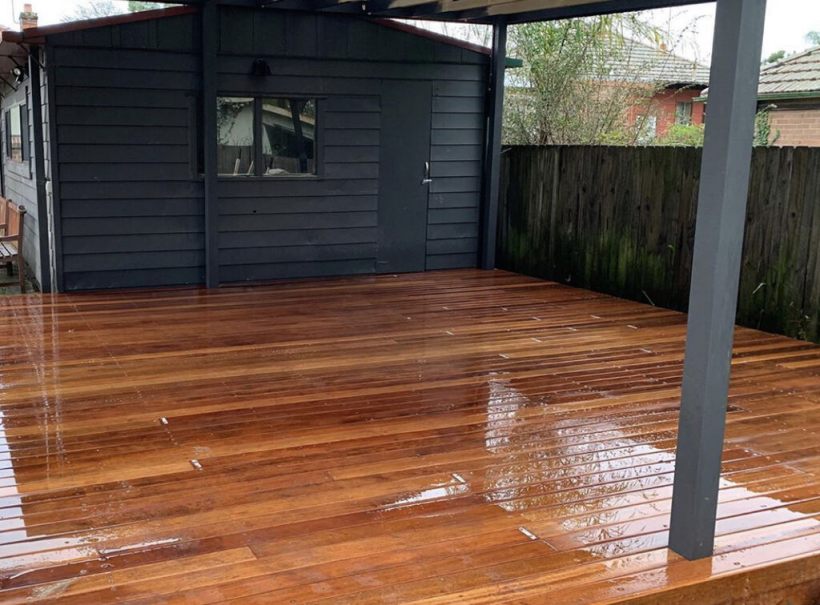 Newly installed wooden flooring on a covered outdoor deck, showcasing a polished and durable finish, with a dark wooden shed in the background.