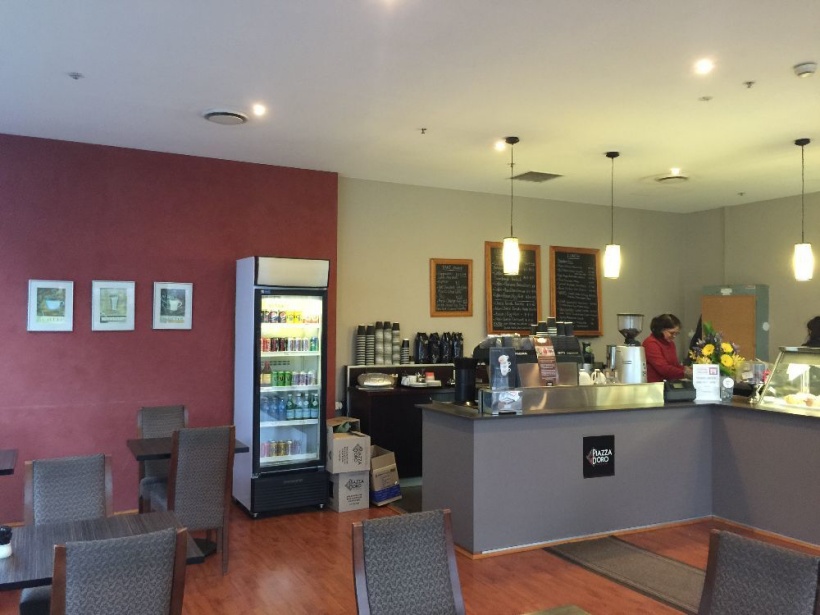 Interior of a commercial cafe featuring a modern shopfitting design with counters, shelves, lighting fixtures, and a refrigerator, creating a functional and visually appealing space.