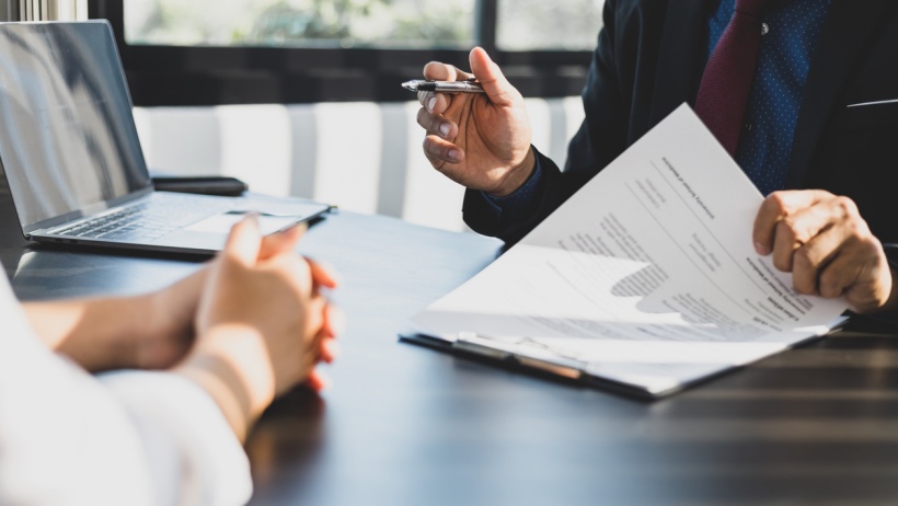 A business professional discussing funding options with a client, holding a document and pen at a desk with a laptop in the background