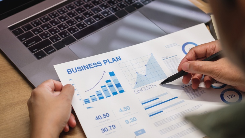 A construction expert reviewing a detailed business plan document with charts and graphs, sitting at a desk with a laptop in the background