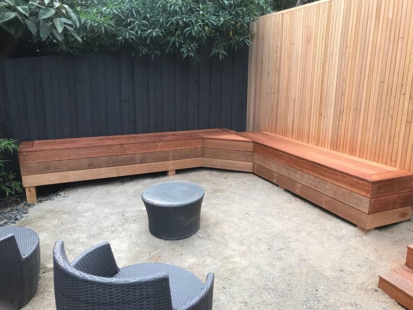 An outdoor custom-built wooden bench seating area with a modern design, set against a wooden privacy fence and surrounded by garden greenery.