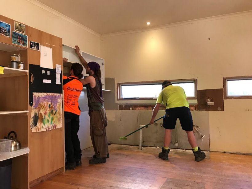 Three carpenters working in an indoor space; two are preparing the wall structure, while one sweeps the floor, showcasing teamwork in a renovation project.